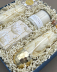 Close angled view of the Cheers to You gift box showing a gold-dipped stemless champagne glass, Belgian chocolate chip cookies in botanical handmade paper wrap, Beverly + 3rd Lavender Sage soy candle, and gold-tipped matches in a corked glass vial, nestled in white crinkle paper fill.