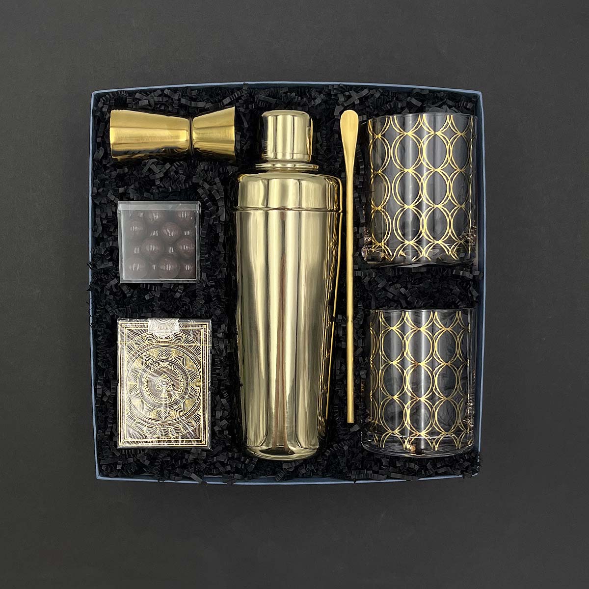 Five O’Clock Gift Box with gold cocktail shaker, lowball glasses, jigger, bar spoon, playing cards, and chocolate caramels