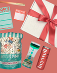 Gift box with a red ribbon, colorful popcorn bag, 'Today's Plan of Attack' notepad, 'High Five' notepad, and two energy drink cans on a pink background.