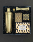 Overhead view of the Happy Hour gift box on a gray background, featuring a gold stainless steel cocktail shaker, gold bar spoon, gold double jigger, Citizen playing cards, Celebrity Trivia game by Games Room, and champagne bubble gummies, nestled in black crinkle paper fill.