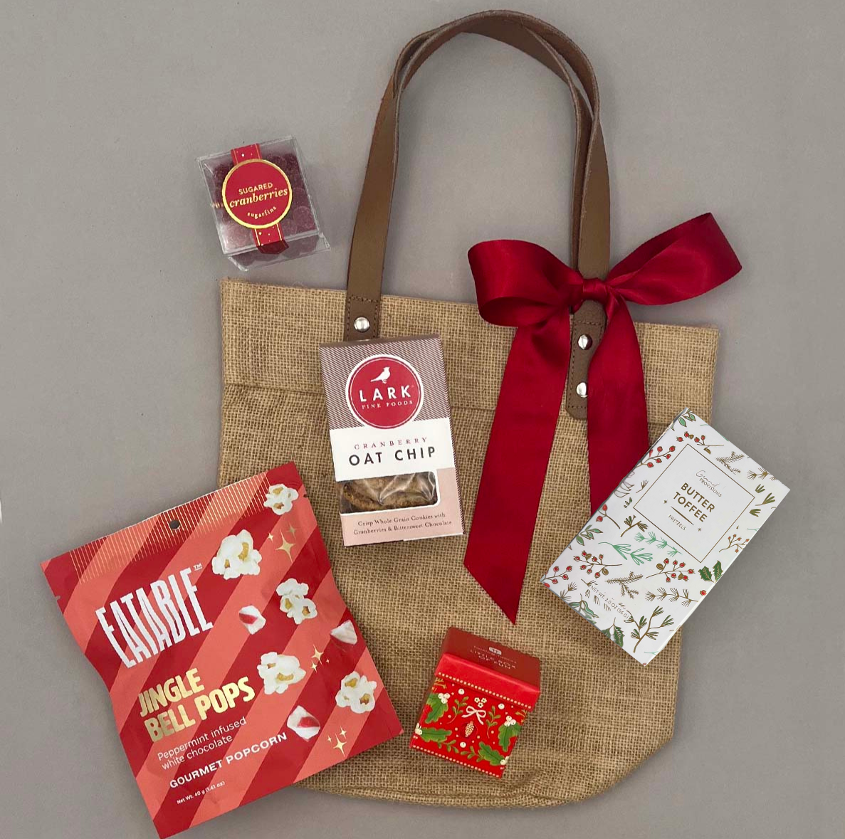 The Holiday Tote by Thoughty — gourmet holiday gift set in a jute tote with leather handles, featuring cookies, popcorn, pretzels, and festive treats.
