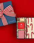 Festive red and green Christmas gift box featuring a candy cane tea towel, striped candle, holiday game, matches, and peppermint candy.