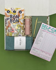 Gift box with Bon Bon candies, a notebook, pen, and self-care tracker on a green background