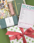 Gift set with self-care items including a pink bottle, green socks, and a self-care weekly tracker on a green surface.