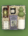 Rise and Shine gift box on a sage green background, overhead view, featuring Wehrloom honey, Berry Best Jam, Sweet Deliverance blueberry granola, a wooden honey dipper, Terre à Terre Verbena soap, a sage green double-gourd bud vase, and a white ceramic tumbler, nestled in white crinkle paper fill.