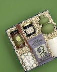 Overhead view of the Rise and Shine gift box featuring small-batch honey, berry jam, blueberry sunflower butter granola, a wooden honey dipper, Provence-made Verbena soap, a sage green ceramic bud vase, and a white porter tumbler nestled in crinkle paper fill.