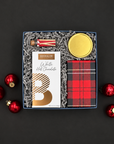 Warm Wishes holiday gift box with white hot chocolate, chocolate chip cookies, Snow & Pine candle, and peppermint-striped matches on a black background with red ornaments.