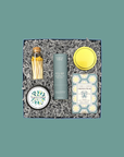 Thoughty Welcome Relief stress relief gift box open beside its navy gift box tied with a teal satin ribbon, showing curated wellness products including Thistle Farms essential oil roll-on, French pear soap, body butter, and gold matches nestled in gray crinkle paper.