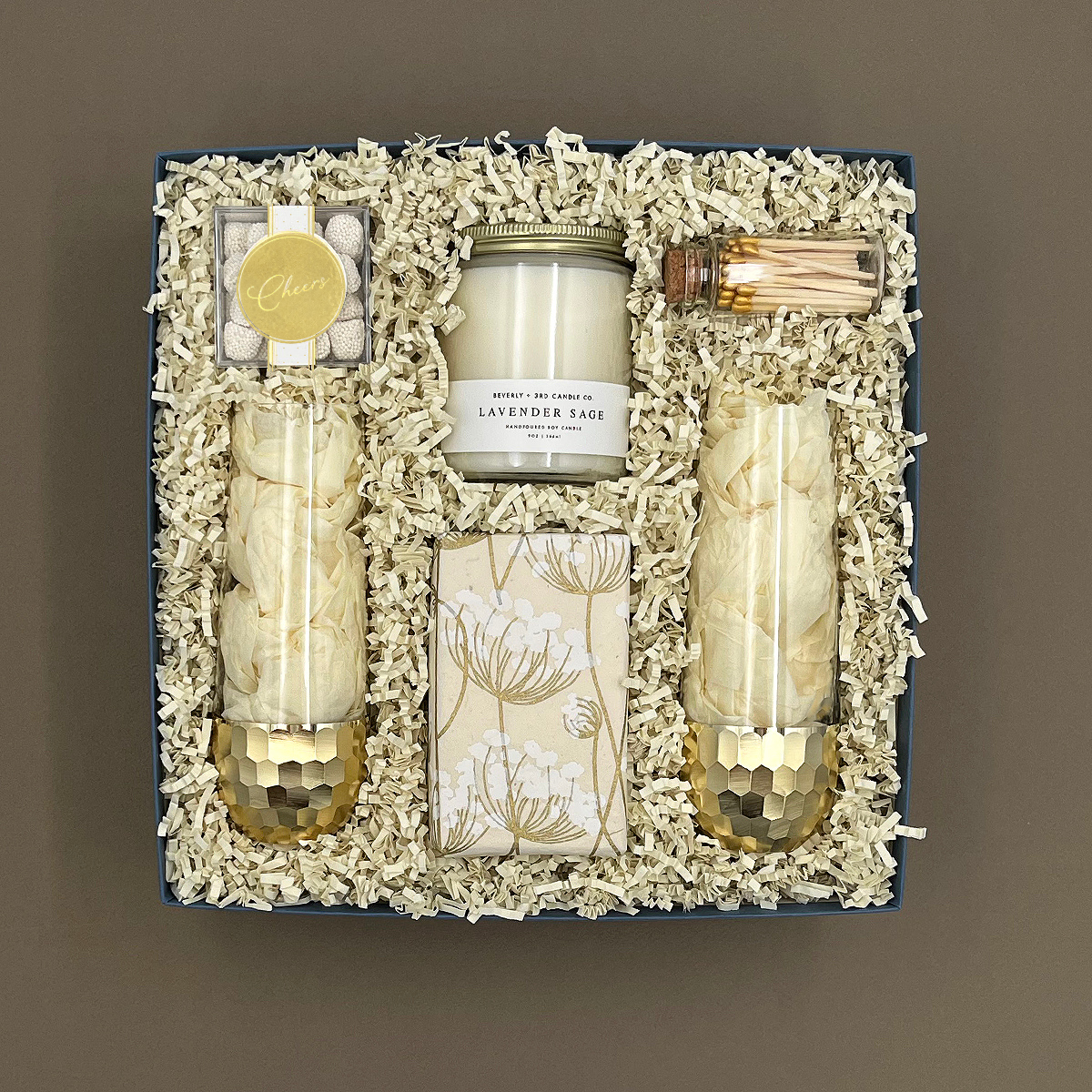 Overhead view of the Cheers to You celebration gift box showing two gold-dipped stemless champagne glasses, Beverly + 3rd Lavender Sage candle, champagne bubble gummies, botanical-wrapped Belgian chocolate chip cookies, and a corked vial of gold-tipped matches in white crinkle paper.