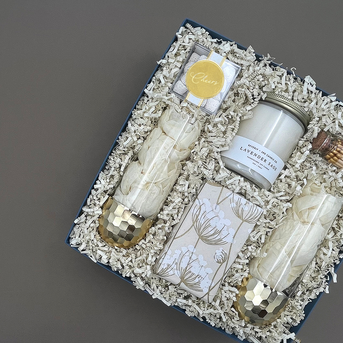 Angled overhead view of the Cheers to You gift box featuring two gold-dipped stemless champagne glasses wrapped in tissue, a Lavender Sage candle, champagne bubble gummies with a gold Cheers seal, botanical-wrapped cookies, and gold-tipped matches in a glass vial.