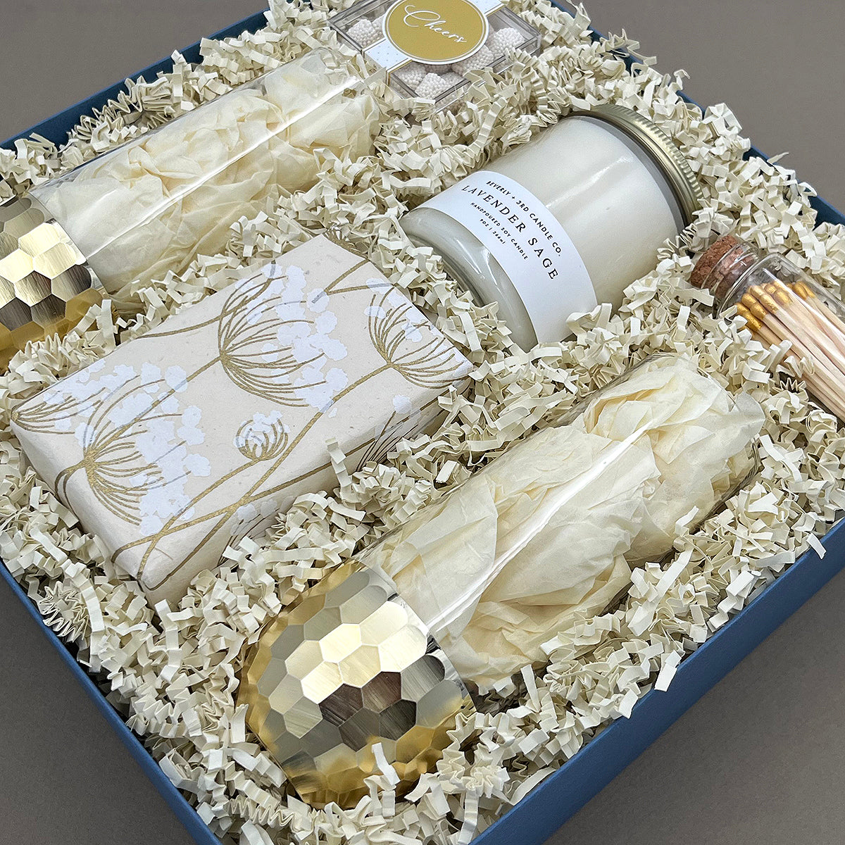 Close angled view of the Cheers to You gift box showing a gold-dipped stemless champagne glass, Belgian chocolate chip cookies in botanical handmade paper wrap, Beverly + 3rd Lavender Sage soy candle, and gold-tipped matches in a corked glass vial, nestled in white crinkle paper fill.