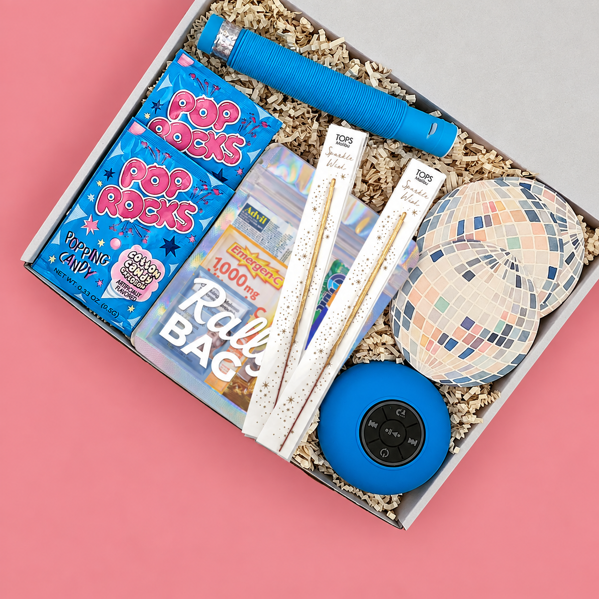 A curated party gift box filled with party supplies, including  a blue portable speaker, disco ball coasters, Pop Rocks candy, and a hangover kit.