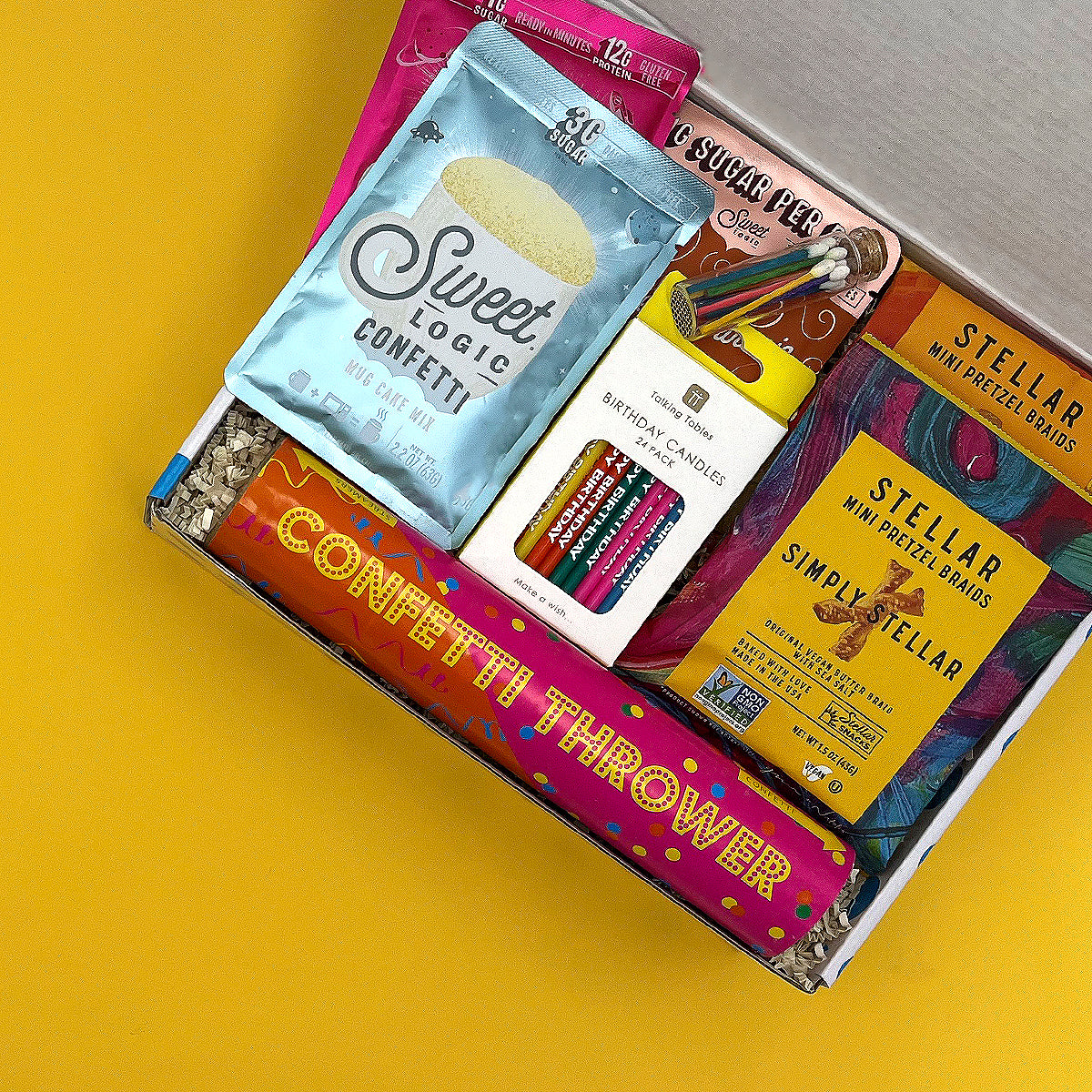 Assorted party supplies including confetti, candles, and mini pretzel bags in a box on a yellow background.