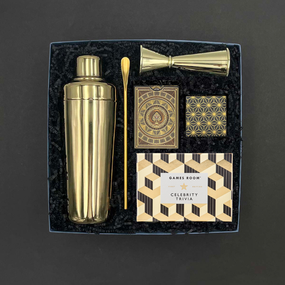 Overhead view of the Happy Hour gift box on a gray background, featuring a gold stainless steel cocktail shaker, gold bar spoon, gold double jigger, Citizen playing cards, Celebrity Trivia game by Games Room, and champagne bubble gummies, nestled in black crinkle paper fill.