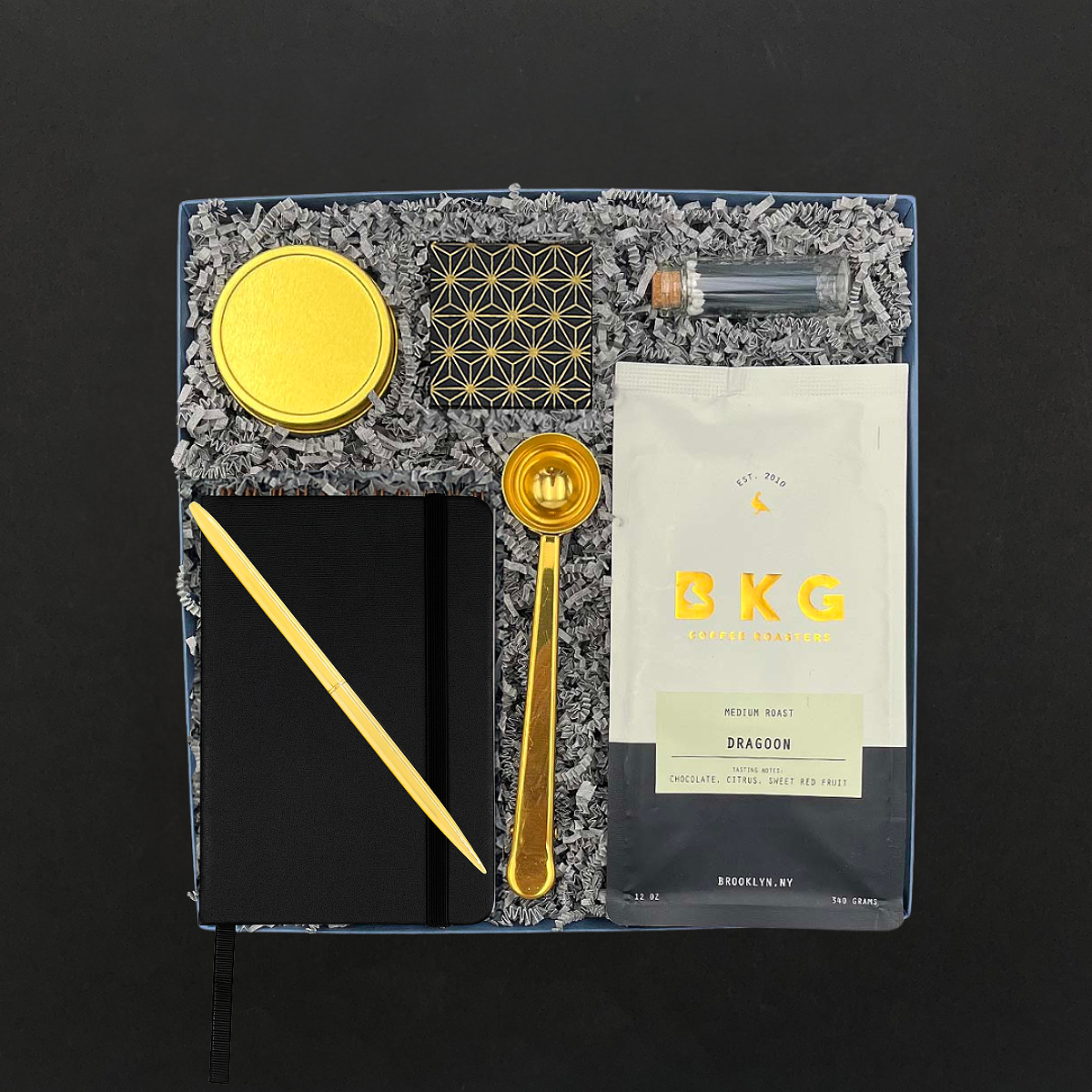 Gift collection including artisan coffee, a gold coffee spoon, candle, matches and a notebook.