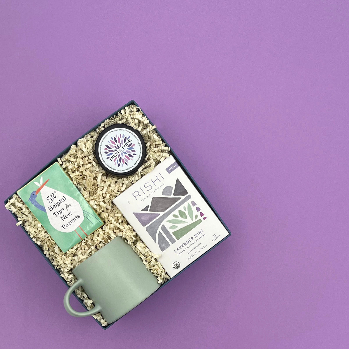 Thoughty Mama to Be pregnancy gift box at an angle, featuring Rishi Lavender Mint Tea, Thistle Farms body butter, 52 Helpful Tips for New Parents card deck, and a sage green ceramic mug nestled in cream crinkle paper against a soft purple background.