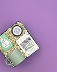 Thoughty Mama to Be pregnancy gift box at an angle, featuring Rishi Lavender Mint Tea, Thistle Farms body butter, 52 Helpful Tips for New Parents card deck, and a sage green ceramic mug nestled in cream crinkle paper against a soft purple background.