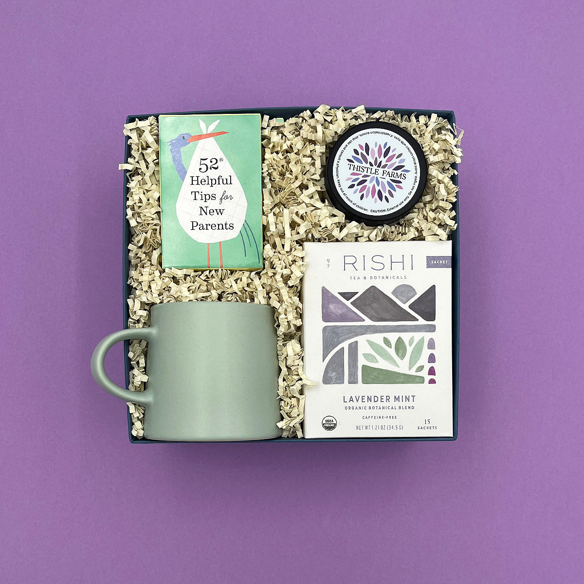 Thoughty Mama to Be baby shower gift box with Rishi Lavender Mint Tea, Thistle Farms body butter, 52 Helpful Tips for New Parents card deck, and a sage green ceramic mug, packed in a navy gift box with cream crinkle paper on a lavender background.