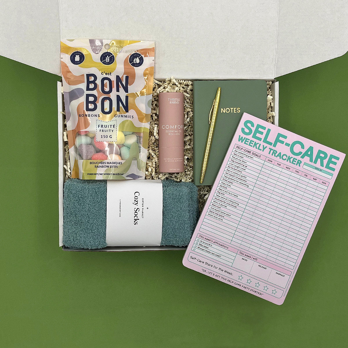 Gift box with Bon Bon candies, a notebook, pen, and self-care tracker on a green background