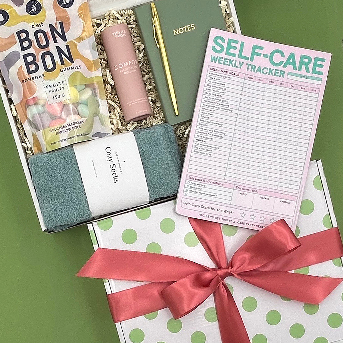 Gift set with self-care items including a pink bottle, green socks, and a self-care weekly tracker on a green surface.