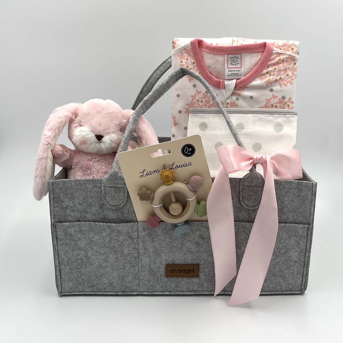 Gray diaper tote with pink ribbon, plush toy, and baby sleep sack, bib and teether rattle on a white background