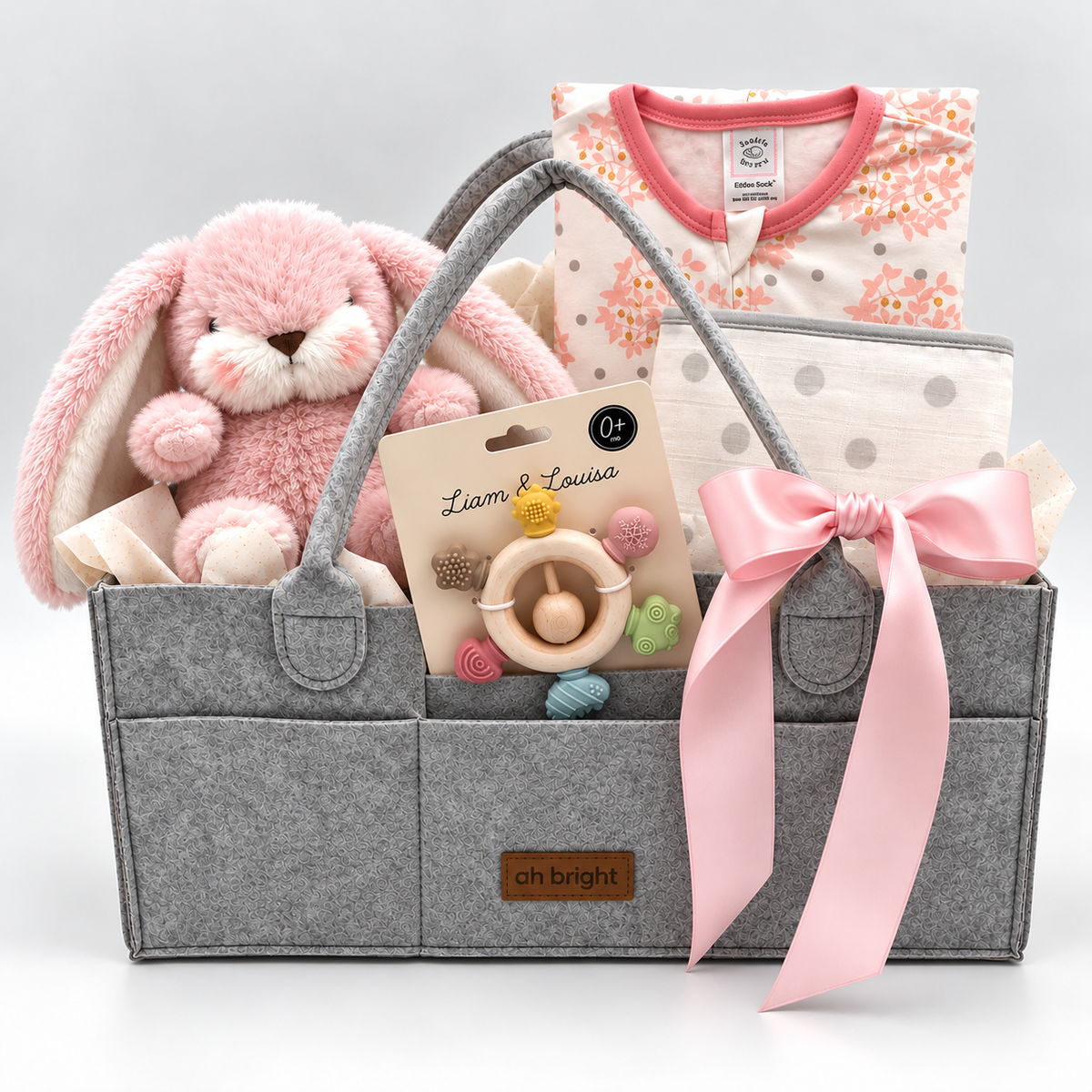 Gray diaper tote with pink ribbon, plush toy, and baby sleep sack, bib and teether rattle on a white background