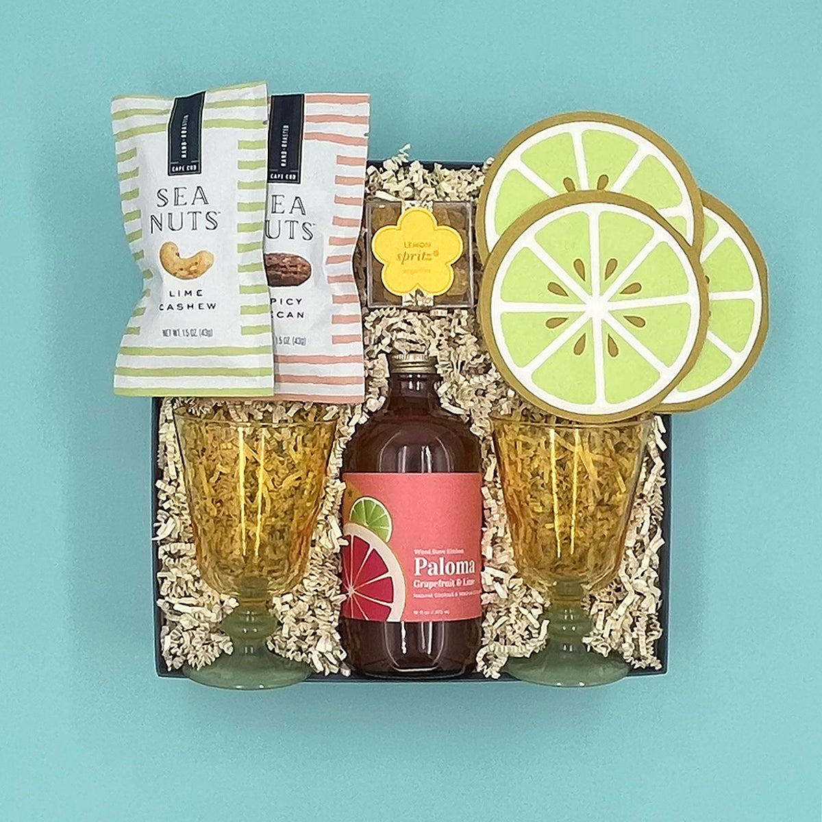 Gift set with Paloma bottle, lime-themed coasters, and sea nut snacks on a blue background