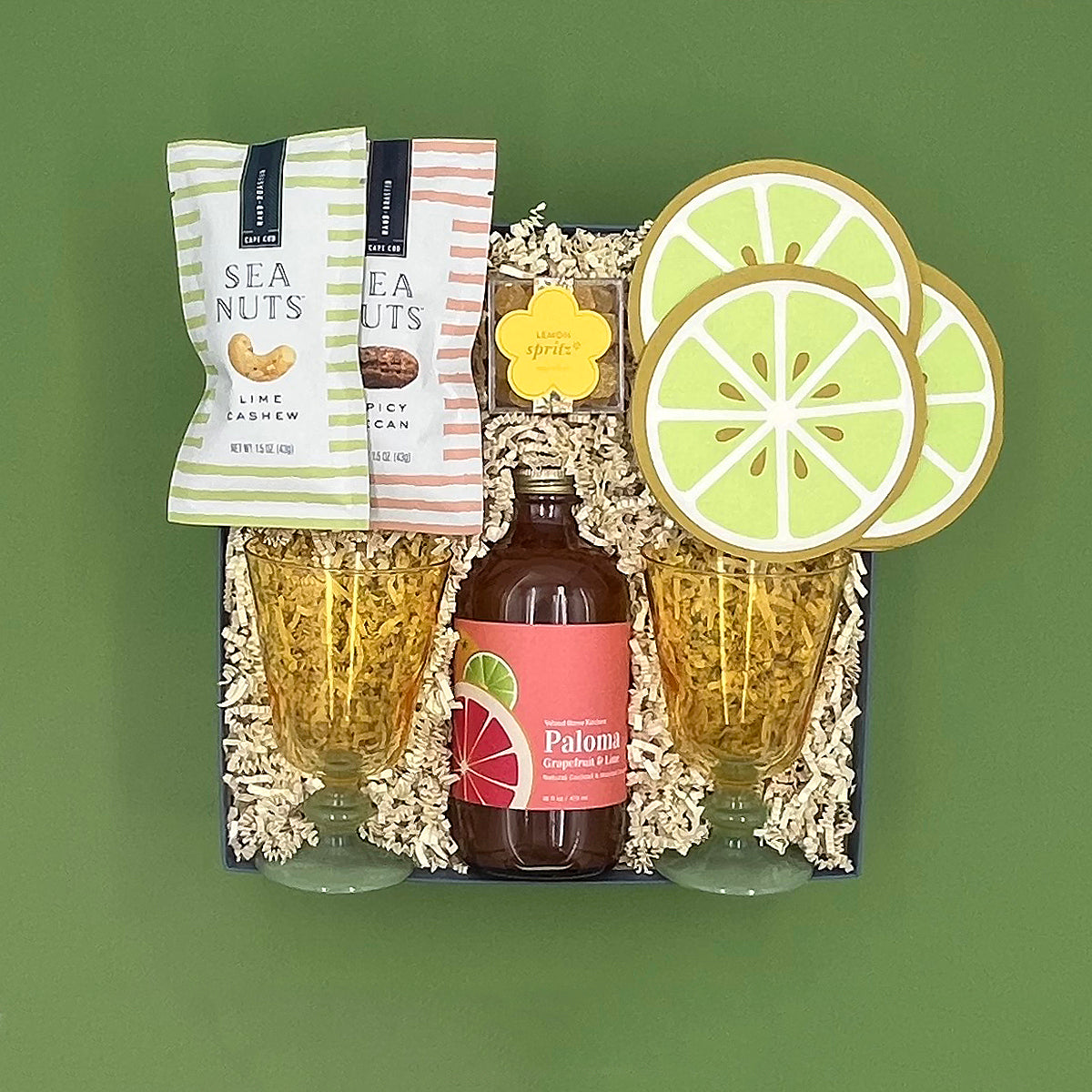 Gift set with Paloma bottle, glasses, and snacks on a green background