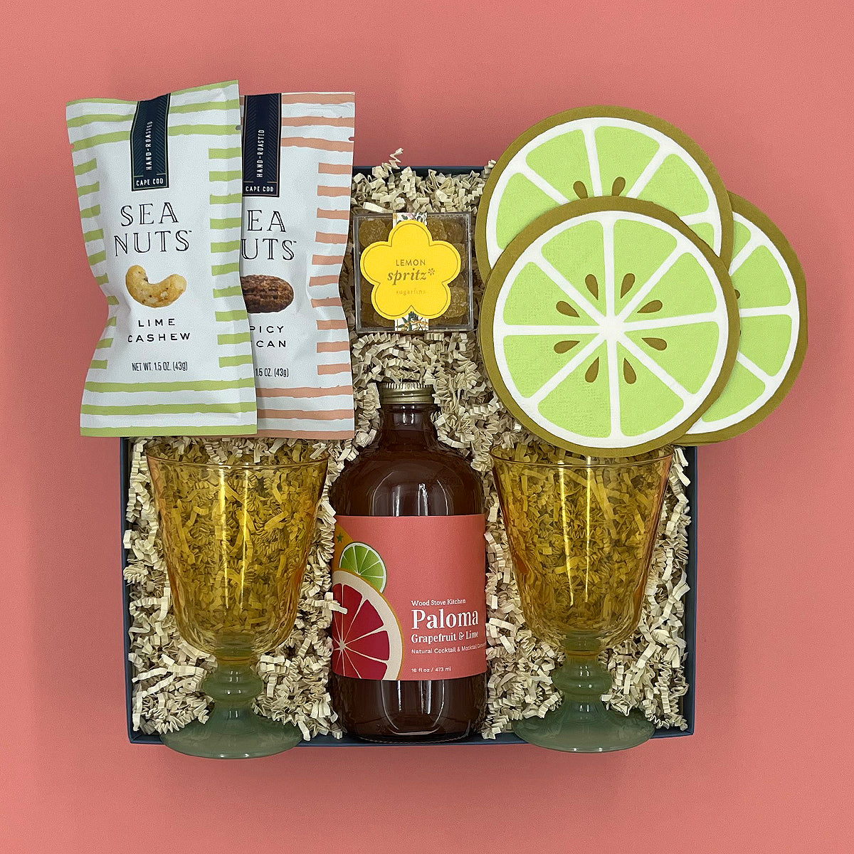 Gift set with Paloma bottle, lime-themed glasses, and sea nut snacks on a pink background