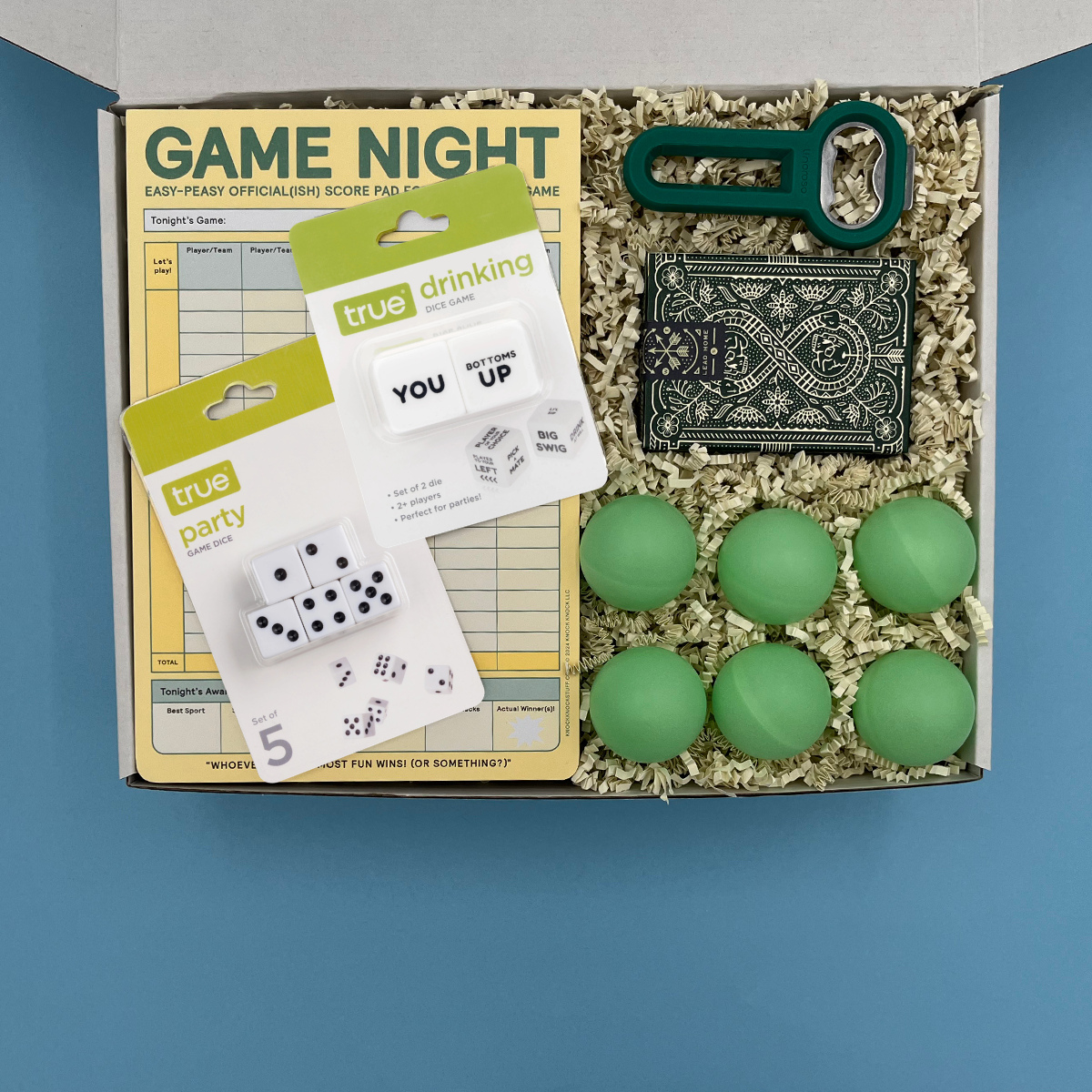 Box containing college game night items including dice, score pads, and green balls on a blue background