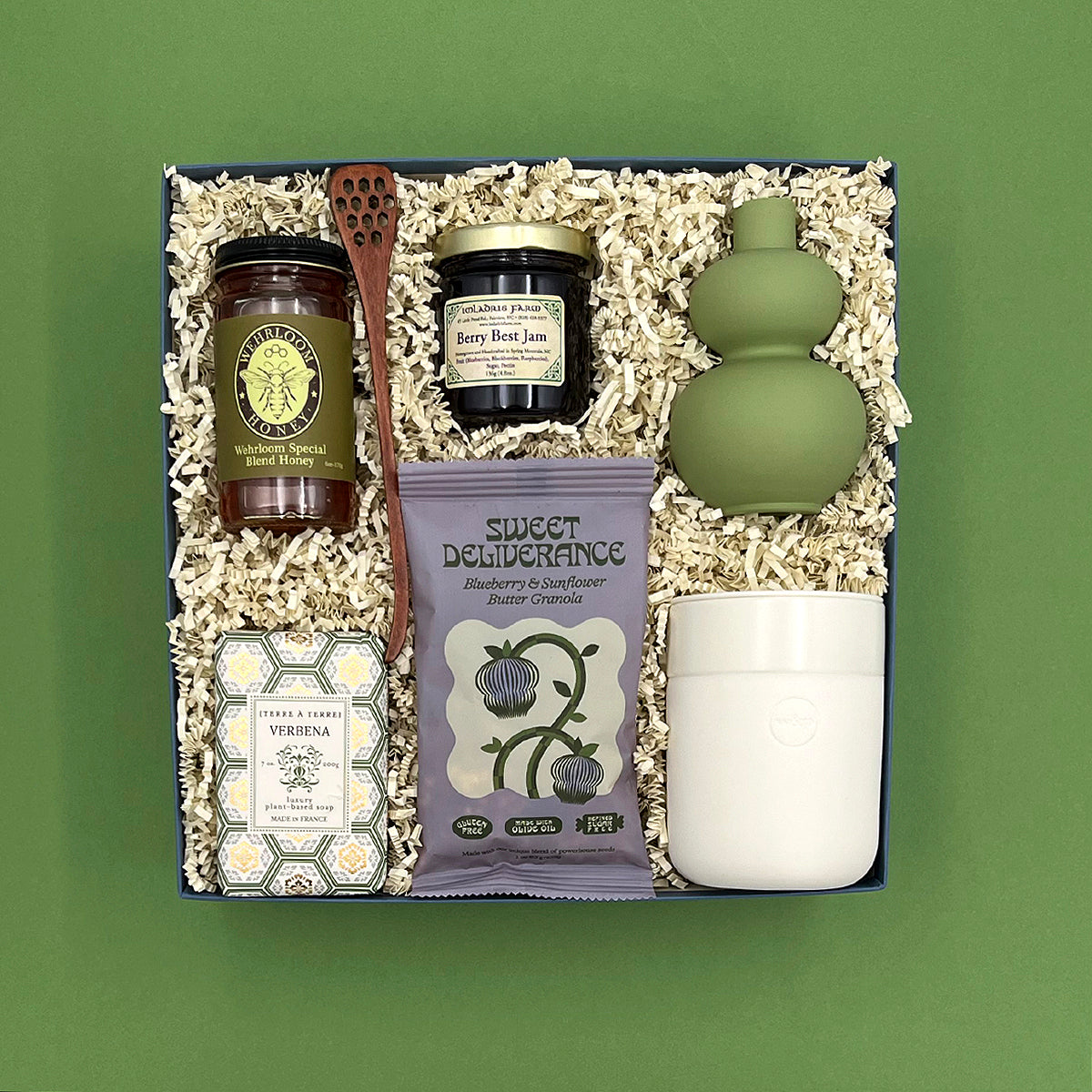 Rise and Shine gift box on a sage green background, overhead view, featuring Wehrloom honey, Berry Best Jam, Sweet Deliverance blueberry granola, a wooden honey dipper, Terre à Terre Verbena soap, a sage green double-gourd bud vase, and a white ceramic tumbler, nestled in white crinkle paper fill.