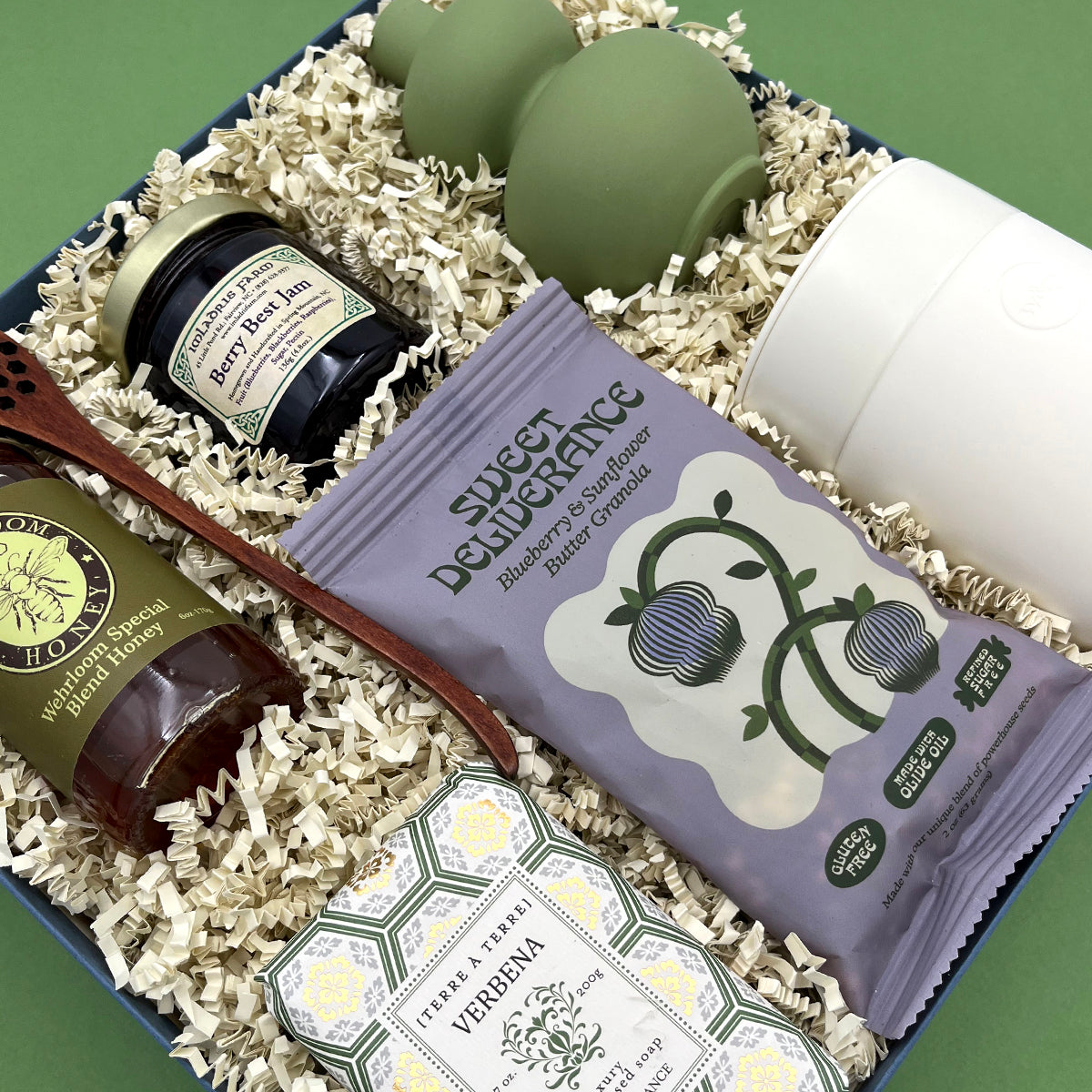 Angled view of the Rise and Shine gift box on a sage green background, showing the Sweet Deliverance granola bag, Berry Best Jam, Wehrloom honey, wooden honey dipper, Verbena soap, white ceramic tumbler, and sage green bud vase packed in white crinkle paper.