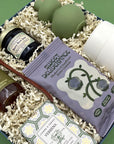 Angled view of the Rise and Shine gift box on a sage green background, showing the Sweet Deliverance granola bag, Berry Best Jam, Wehrloom honey, wooden honey dipper, Verbena soap, white ceramic tumbler, and sage green bud vase packed in white crinkle paper.
