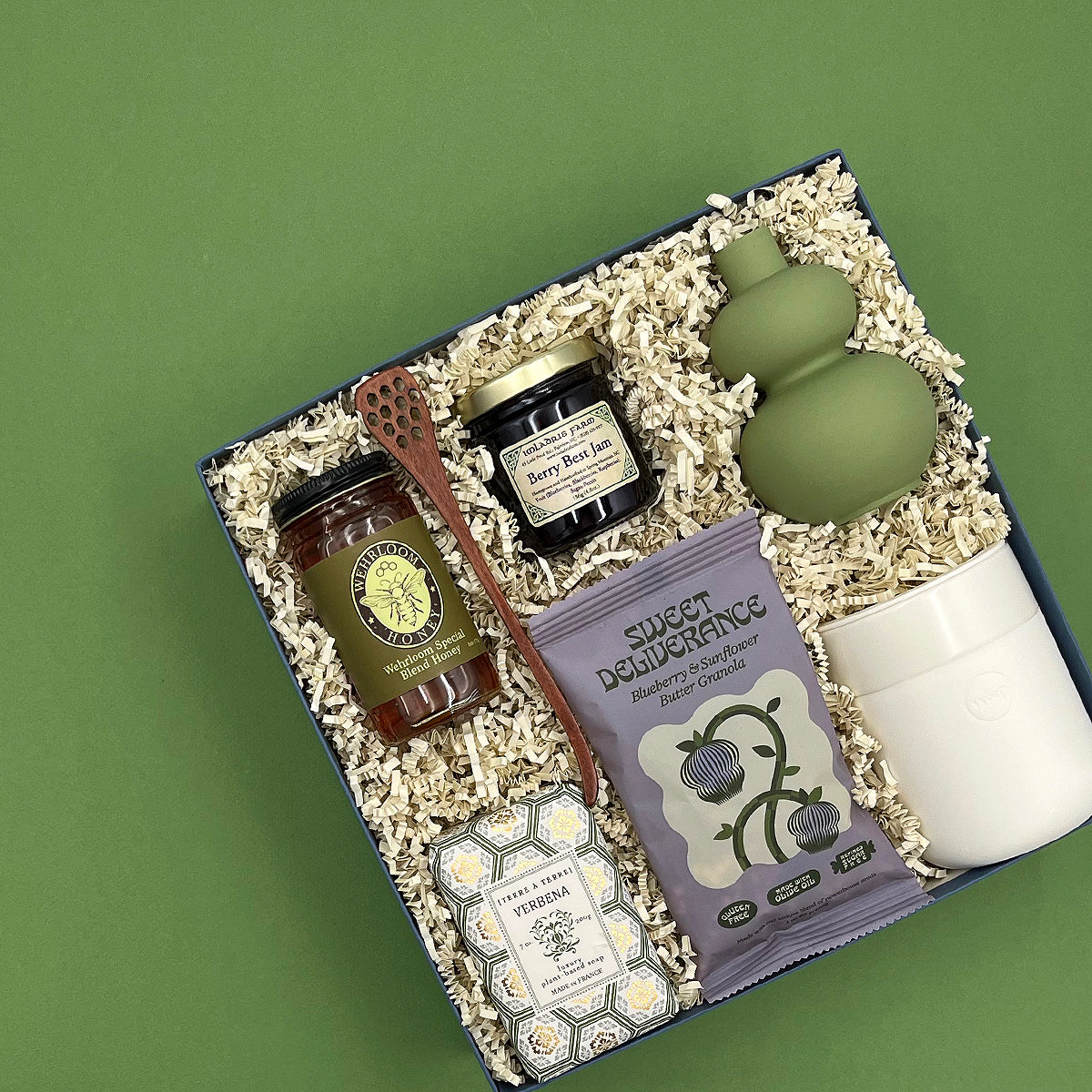 Overhead view of the Rise and Shine gift box featuring small-batch honey, berry jam, blueberry sunflower butter granola, a wooden honey dipper, Provence-made Verbena soap, a sage green ceramic bud vase, and a white porter tumbler nestled in crinkle paper fill.