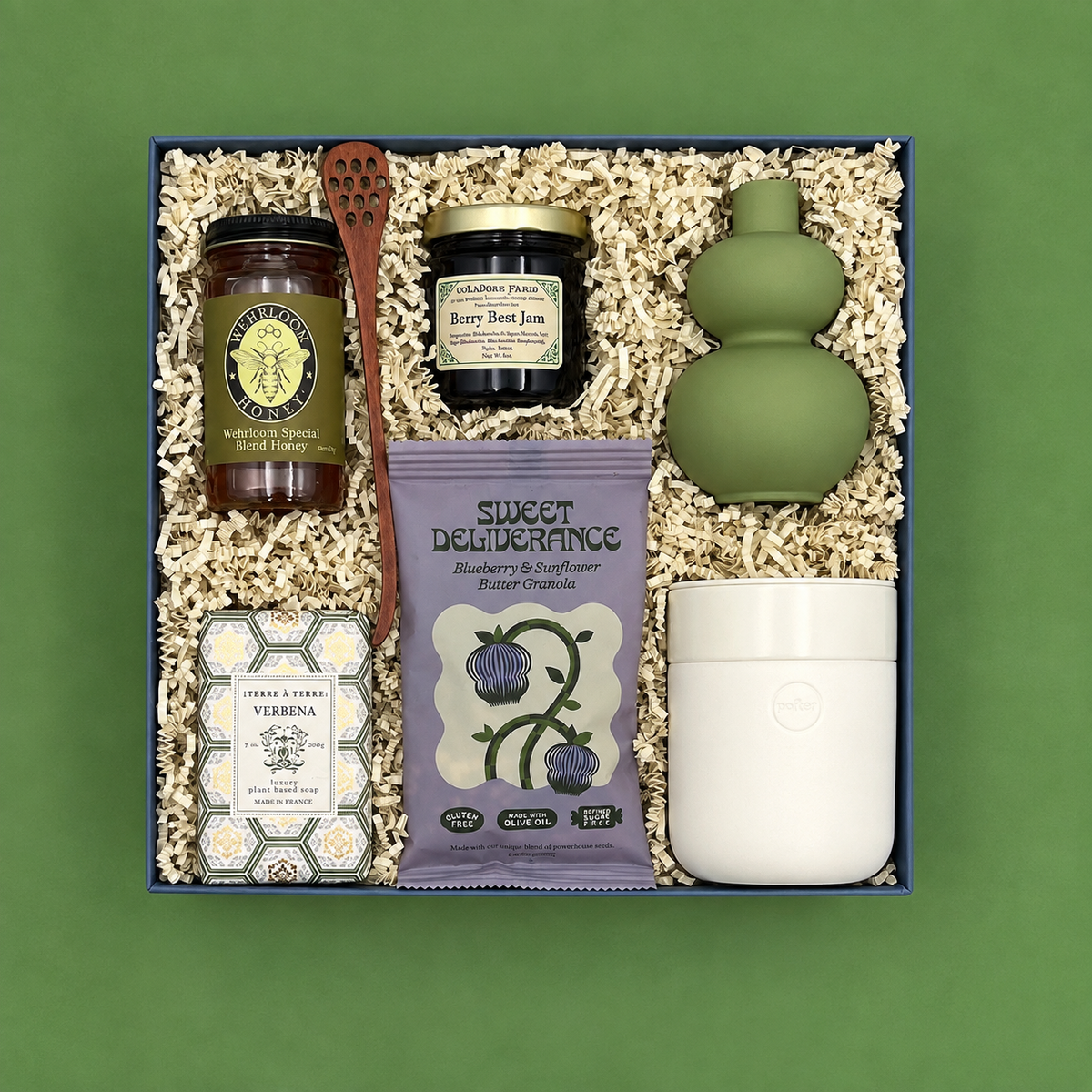 Rise and Shine gift box on a sage green background, overhead view, featuring Wehrloom honey, Berry Best Jam, Sweet Deliverance blueberry granola, a wooden honey dipper, Terre à Terre Verbena soap, a sage green double-gourd bud vase, and a white ceramic tumbler, nestled in white crinkle paper fill.