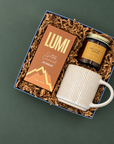 A curated gift box set on a dark green background, featuring a box of Salted Caramel Hot Chocolate, a Vetiver Tonka Candle, and a white ribbed ceramic mug.
