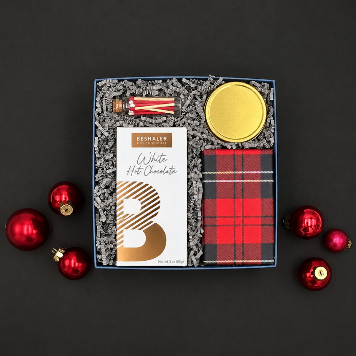 Warm Wishes holiday gift box with white hot chocolate, chocolate chip cookies, Snow & Pine candle, and peppermint-striped matches on a black background with red ornaments.