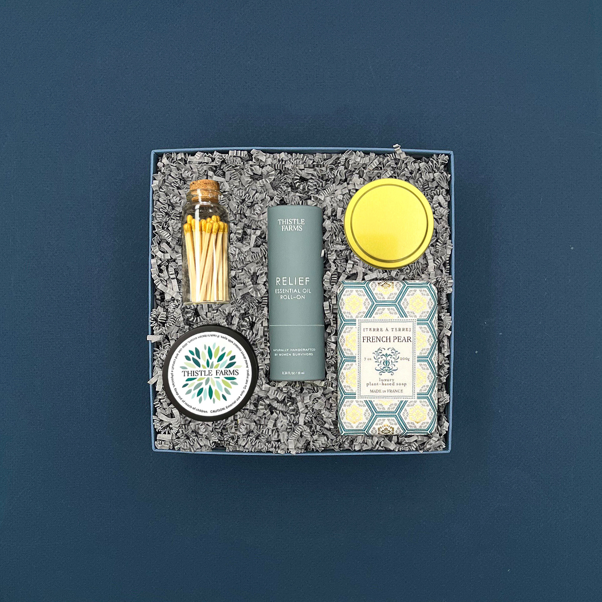 Thoughty Welcome Relief wellness gift box with Thistle Farms essential oil roll-on, French pear soap, eucalyptus mint body butter, gold matches in a corked vial, and a eucalyptus jasmine candle, nestled in gray crinkle paper against a deep navy background.