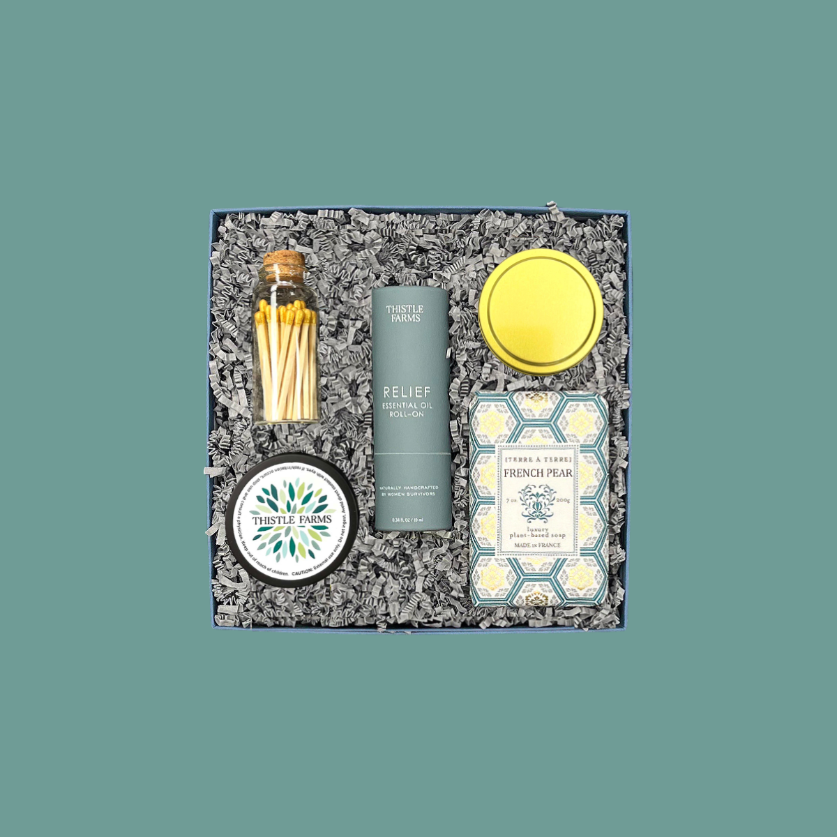 Thoughty Welcome Relief stress relief gift box open beside its navy gift box tied with a teal satin ribbon, showing curated wellness products including Thistle Farms essential oil roll-on, French pear soap, body butter, and gold matches nestled in gray crinkle paper.