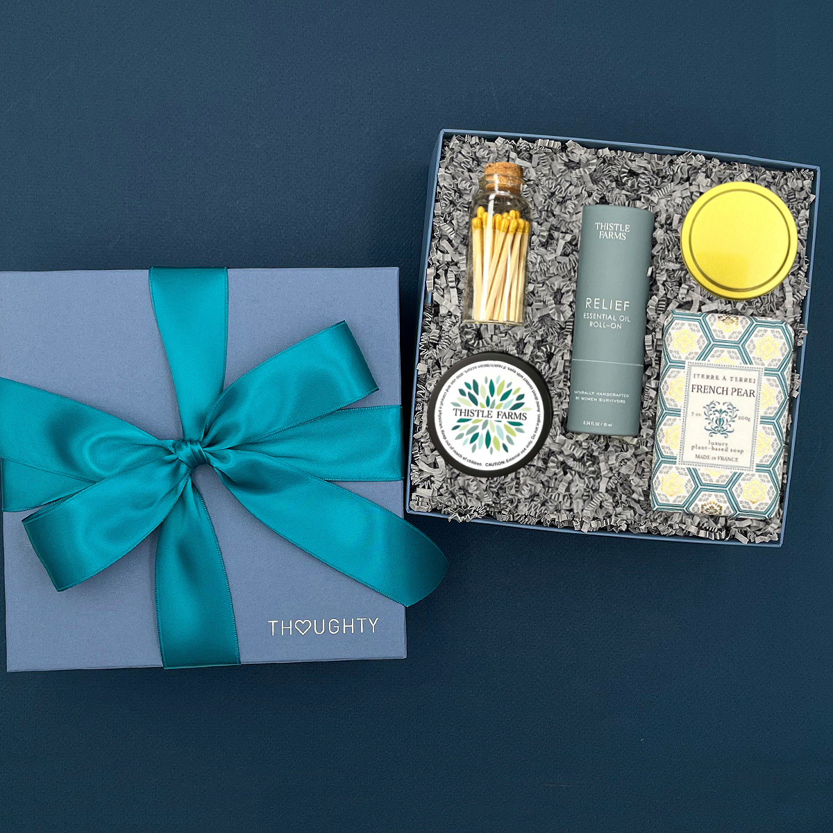 Thoughty Welcome Relief self-care gift box displayed against a soft teal background, featuring Thistle Farms Relief essential oil roll-on, Terre a Terre French pear soap, eucalyptus mint body butter tin, and gold matches in a corked glass vial.