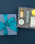 Thoughty Welcome Relief self-care gift box displayed against a soft teal background, featuring Thistle Farms Relief essential oil roll-on, Terre a Terre French pear soap, eucalyptus mint body butter tin, and gold matches in a corked glass vial.